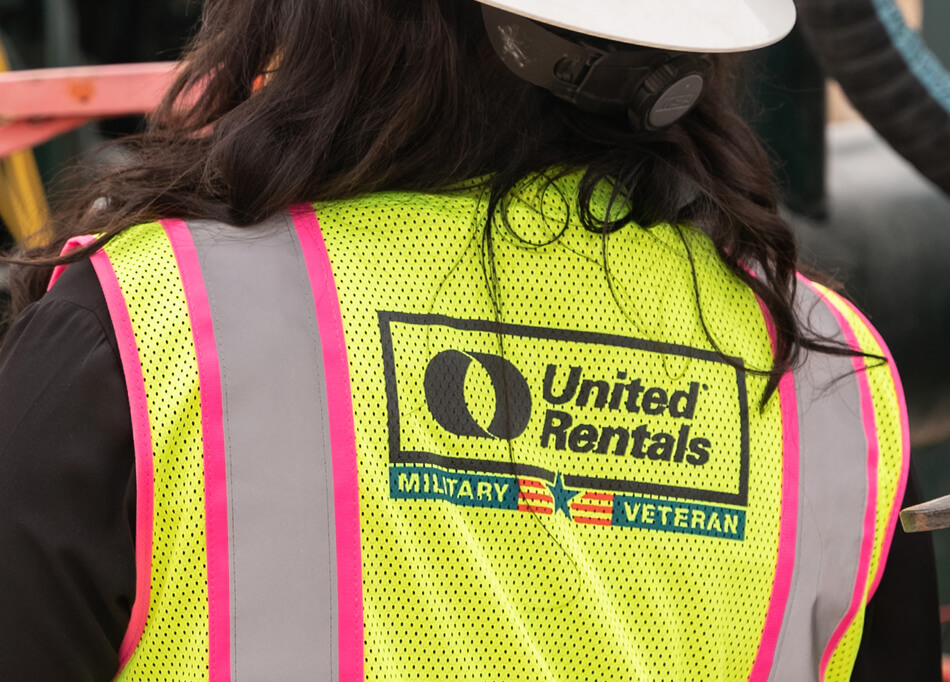 The back of a female United Rentals employee in a yellow vest with a military veteran designation.